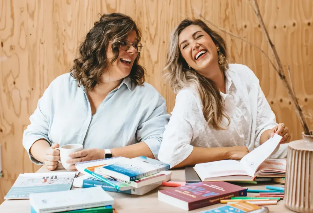 Twee ergotherapeuten lachen samen aan een tafel met boeken